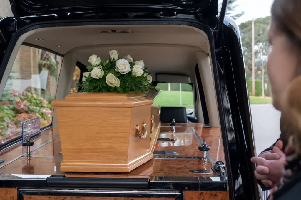 Cercueil en bois clair orné de roses blanches dans un corbillard.